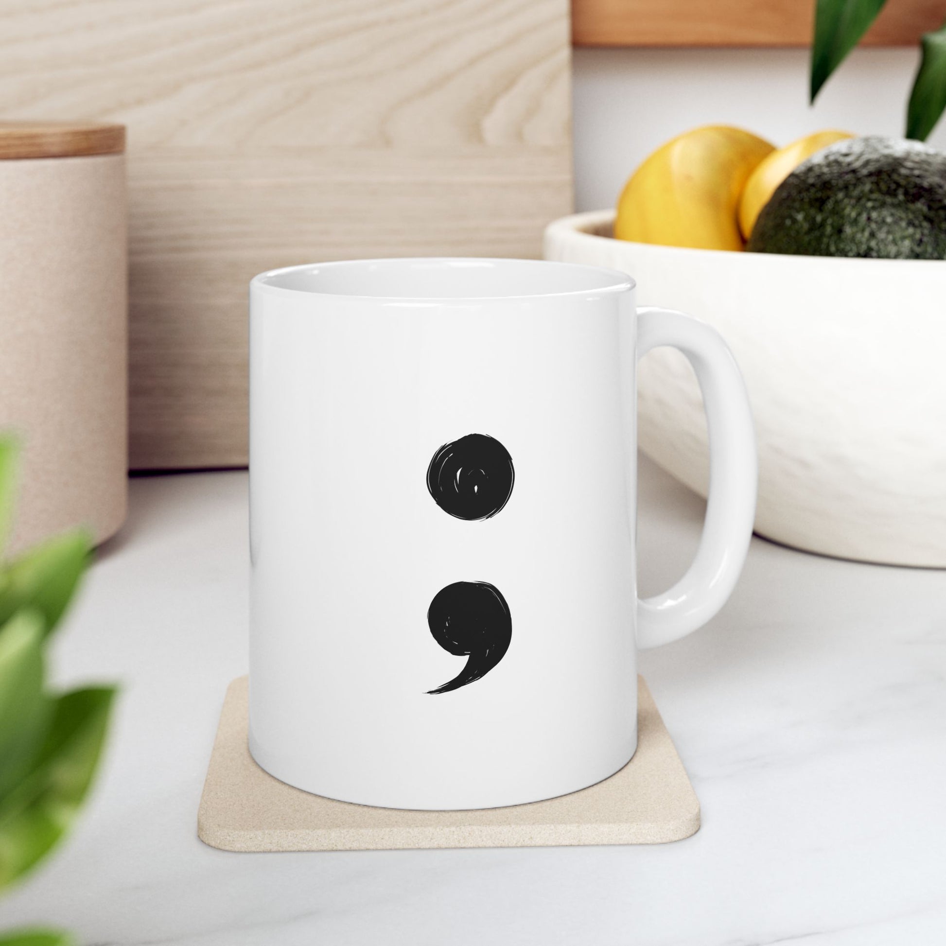 White mug with a black semicolon design on a coaster, placed on a light surface with a plant and container in the background.