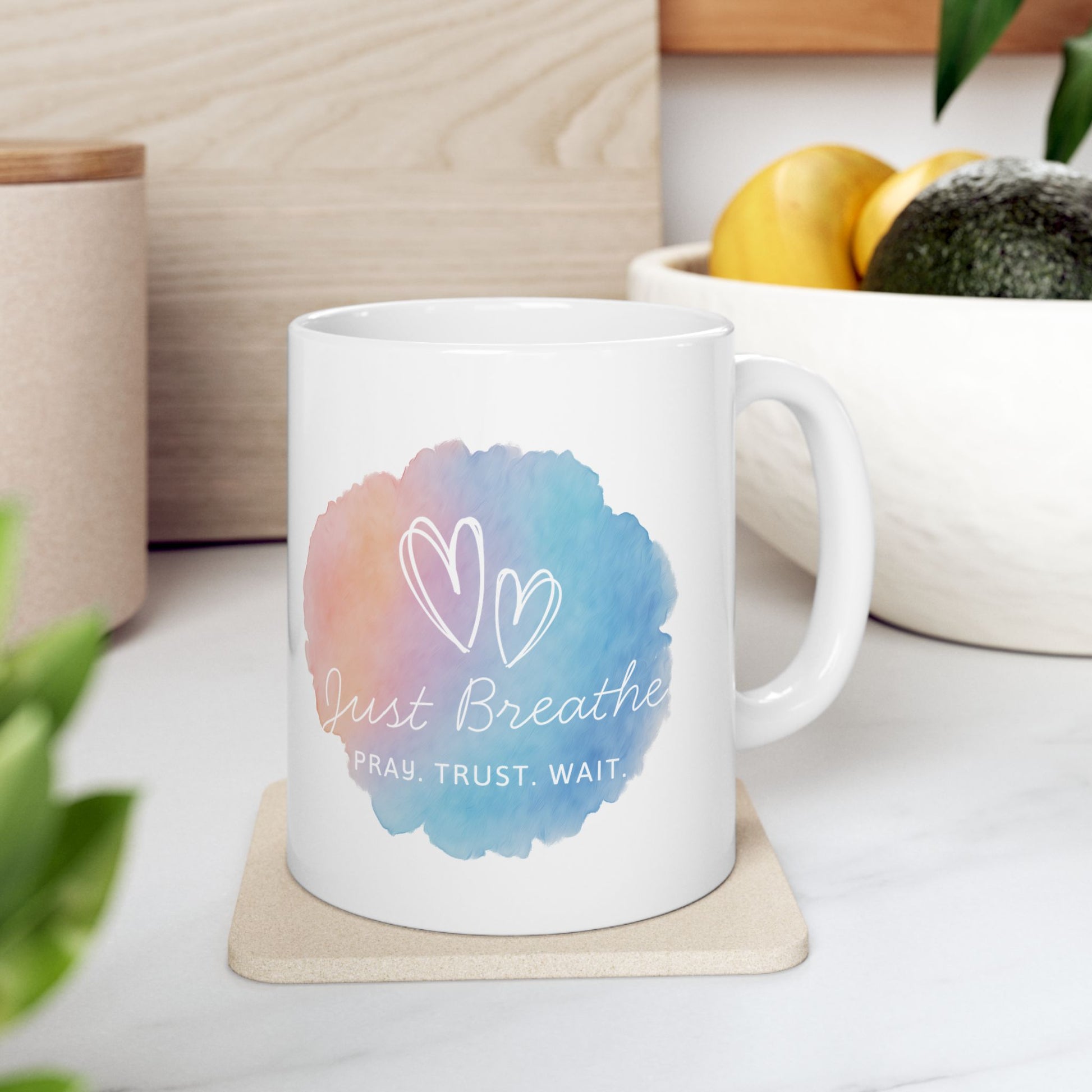 White mug with colorful design and text on a white surface with plants and a plant pot in the background